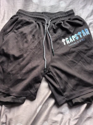 Svarta shorts med tryck och snörning - Svarta shorts med vit och blå text på vänster sida, elastisk midja och snörning framtill. Tillverkade i ett syntetmaterial som känns sportigt och bekvämt. Perfekta för en avslappnad streetwear-look.