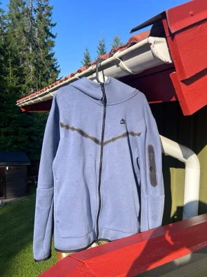 Nike Tech Fleece Hoodie (Blå) - Säljer en ljusblå Nike Tech Fleece zip hoodie i storlek M. Skön och stilren modell med svarta detaljer och ficka på ärmen. Perfekt till vardags. Bra använt skick med normalt slitage. Se bilder för skick.