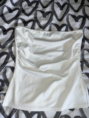Tubtopp - Säljer en stilren vit bandeau tubtopp med draperad framsida. Toppen är axelbandslös och har en mjuk, stretchig känsla som gör den bekväm att bära. Perfekt att matcha med jeans eller shorts/kjol för en clean look. Någon typ av liten reva finns