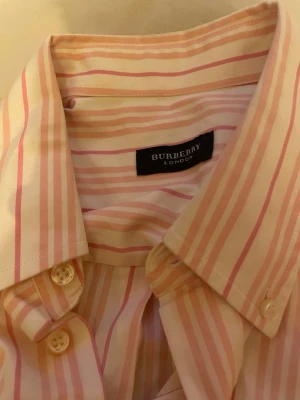 Randig skjorta från Burberry - Säljer en klassisk skjorta från Burberry med rosa och vita ränder. Skjortan har lång ärm, knappar framtill och en bröstficka. Tillverkad i mjuk bomull och har en avslappnad passform. Perfekt för dig som gillar stilrena och tidlösa plagg.