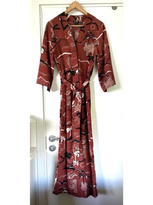 Monki - Mönstrad rödbrun jumpsuit med knytskärp - storlek S - Superfin brun / rostbrun / roströd jumpsuit / onepiece från MONKI i storlek S med ett unikt mönster av vita och svarta palmer, berg och djur. Tidlös omlottdesign upptill med V-ringning och en knytband i midjan som framhäver figuren. Ärmarna är trekvartslånga och vida, vilket ger en avslappnad känsla. Luftiga vida och croppade byxben och diskreta fickor (yay!) i sidan. Enkel att styla till både vardag och fest. Mycket gott skick, som ny!