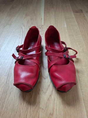 Trippen, handmade LOAFERS/BALLERINA/MOCKASIN strlk 39 i rött läder med remmar - Trippen modell Travel, handmade, välgjorda i ganska tjockt, men mjukt följsamt, ekologiskt läder och sula i svart naturgummi. En ganska lätt sko med avslappnad känsla. Den röda färgen drar varken åt blått eller gult. Köpta för ca 12 år sedan och är använda kanske fem gånger. Några små mörkare partier i lädret mot hälen. Annars är de minimalt slitna. Med vaxning en gång om året kommer de att hålla vår, sommar som höst.