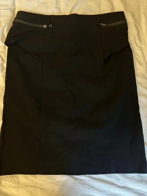 Svart pennkjol med dragkedjedetalj - Stilren svart pennkjol med markerad midja och coola dragkedjor framtill. Kjolen har en klassisk rak passform och är perfekt för en trendig och enkel look. Materialet känns mjukt och följsamt, troligen polyester eller bomullsmix. Ingen storlek står, men passar som en S/M