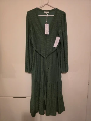 Grön långklänning med broderade detaljer - Snygg grön långklänning med v-ringning, broderade hålmönster och volang nedtill. Klänningen har lång ärm och ett knytband i midjan för en smickrande passform. Perfekt för dig som gillar romantiska detaljer och en avslappnad men chic stil.
