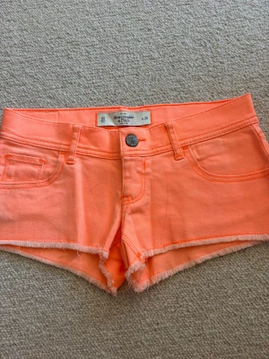 Korall shorts från Abercrombie & Fitch - Snygga korall shorts från Abercrombie & Fitch med fransig kant och klassisk femficksdesign. Shortsen har knapp och dragkedja framtill samt bakfickor med dekorativ söm. Perfekta för varma dagar.💓