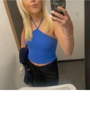 Blå  halterneck topp - Säljer en snygg blå croppad halterneck topp med smala axelband som korsas i nacken. Toppen är ärmlös och har en tight passform, perfekt till jeans eller kjol. Materialet känns mjukt och stretchigt, troligen polyester eller bomull.