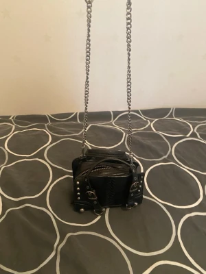 Svart axelväska med kedjerem från Zara - Cool svart axelväska i konstläder med silverfärgade nitar, spännen och detaljer. Väskan har en lång kedjerem och dekorativa sömmar framtill. Perfekt för dig som gillar en edgy och alternativ stil.