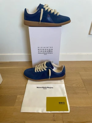 Maison Margiela gats blå - Snygga blå sneakers från Maison Martin Margiela med beigea skosnören och klassisk gummisula. Ovandelen är i mocka och läder, vilket ger en lyxig känsla. Perfekta för dig som vill ha en stilren och trendig look.