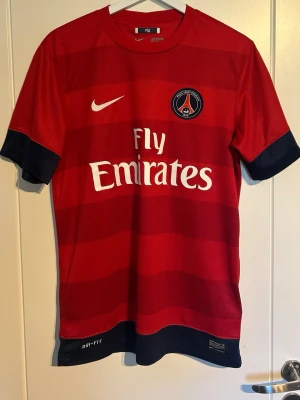 Röd PSG matchtröja från Nike 2012/2013 - Röd PSG matchtröja med mörkblå detaljer och vita loggor. Tröjan har Paris Saint-Germain klubbmärke på bröstet, Nike-logga och Fly Emirates-tryck. Tillverkad i Dri-FIT material som andas, perfekt för fotbollsträning eller match. Beckham nummer 7 på ryggen. Trycket lite förstört dock. Priset kan diskuteras 