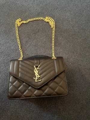 Svart quiltad YSL handväska - Snygg svart quiltad handväska från YSL med guldfärgad kedja och ikonisk YSL-logga framtill. Väskan har ett elegant kuvertlock och stilrena sömmar som ger en lyxig känsla. Perfekt accessoar för att lyfta vilken outfit som helst.