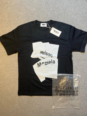 Svart Maison Margiela t-shirt - Säljer en svart t-shirt från Maison Margiela med ett stort vitt grafiskt tryck och logga på bröstet. T-shirten har rund hals och korta ärmar. Perfekt för dig som gillar statement-plagg och vill sticka ut lite extra.