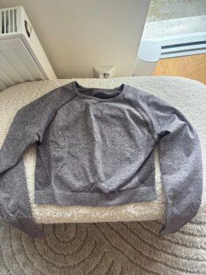 Grå långärmad crop top - Snygg grå långärmad crop top i stretchigt funktionsmaterial. Tröjan har meshdetaljer på sidorna för extra ventilation och en tight passform som passar perfekt till träning eller gym. Rund halsringning och sömlös design för maximal komfort.