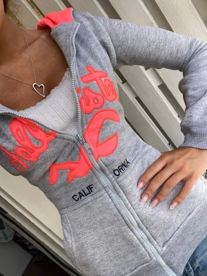 Ljusgrå Hollister zip hoodie med korall rosa broderi - Gullig ljusgrå Hollister zipup hoodie med neon ish korall/ orangerosa broderad text över bröstet, strl M (omsydd till xxs) följ för fler liknande hollister/ abercrombie hoodies!