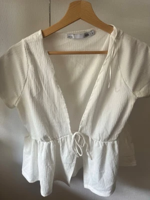 Vit omlottopp med knyt och volang - Säljer en söt vit omlottopp med korta ärmar, knyt i midjan och volang nedtill. Toppen har en djup v-ringning och är gjord i ett ribbat tyg som ger en snygg struktur. Perfekt för dig som gillar en feminin och avslappnad stil.