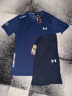 Blått träningsset från Under Armour - Snyggt träningsset med t-shirt och shorts från Under Armour i mörkblått. T-shirten har logga på bröstet och färgglad text på sidan, medan shortsen har dragsko i midjan och logga på benet. Perfekt för gym eller löpning.