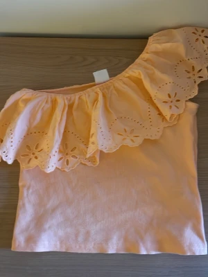 Persikofärgad oneshoulder topp med volang - Söt persikofärgad oneshoulder topp med bred volangkant dekorerad med broderade blommor. Toppen är ärmlös och har en ribbad struktur i tyget. Perfekt för dig som vill sticka ut med en unik detalj.