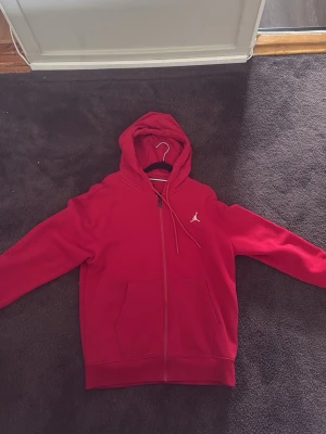 Röd Jordan hoodie med dragkedja - Snygg röd hoodie från Jordan med hel dragkedja och klassisk logga på bröstet. Hoodien har huva med dragsko och två stora fickor framtill. Perfekt för dig som gillar sportig och avslappnad stil.