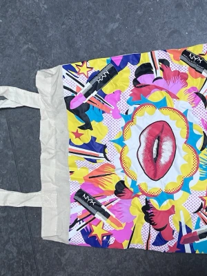Färgglad shoppingväska från NYX - Säljer en färgglad shoppingväska från NYX med pop art-inspirerat tryck av läppar, läppstift och färgsprakande mönster i gult, rosa, blått och svart. Väskan har två långa handtag och är tillverkad i bomull. Perfekt för att bära med sig allt man behöver.