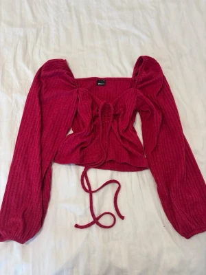 Ribbad rosa/röd topp från Gina Tricot - Trendig ribbad topp från Gina Tricot i en stark röd färg. Modellen har långa puffärmar, croppad passform och snygg snörning framtill som ger en unik look. Perfekt för dig som vill sticka ut lite extra.