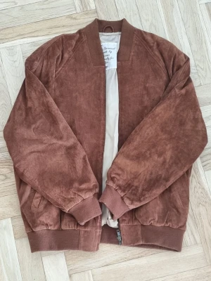 Brun bomberjacka i manchester - Säljer en snygg brun bomberjacka från H&M i  mocka. Jackan har ribbade muddar vid ärmslut och nederkant, samt två sidofickor. Perfekt för dig som vill ha en stilren och avslappnad look.