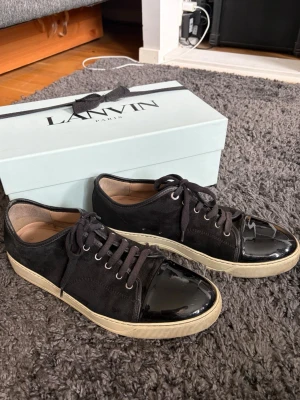 Svarta Lanvin sneakers med lackdetalj - Snygga svarta sneakers från Lanvin med ovandel i mocka och glansig lackad tå. Skorna har svarta skosnören, beige sula och klassisk låg profil. Perfekta för dig som vill ha en stilren och exklusiv look med en twist.