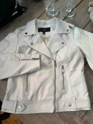 Vit/cremé skinnjacka från Vero Moda - Snygg vit/creméfärgad skinnjacka från Vero Moda med klassisk bikerkrage, dragkedjor på ärmar och framficka samt dekorativa detaljer vid axlar och midja. Jackan har två dragkedjefickor framtill och är helfodrad i ljust tyg. Oanvänd.