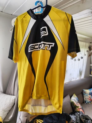 Gul cykeltröja från Scott - Snygg gul cykeltröja från Scott med svart och vitt grafiskt mönster och dragkedja framtill. Tröjan har korta ärmar och är tillverkad i ett lätt och ventilerande material som passar perfekt för cykling. Liten i storlek är mer som l-xl 