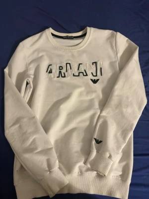 Vit sweatshirt från Armani - Snygg vit sweatshirt från Armani med svart broderad logga och text framtill. Tröjan har rund halsringning, långa ärmar och ribbade muddar vid ärmslut och nederkant. Perfekt för en stilren och trendig look.