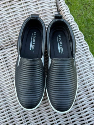 Svarta slip-on sneakers från Skechers - Stilrena svarta slip-on sneakers från Skechers Street med ribbad ovandel i syntetmaterial och vit tjock sula. Skorna har elastiska paneler på sidorna för enkel påtagning och en modern, clean look. Perfekta för dig som gillar minimalistisk stil.