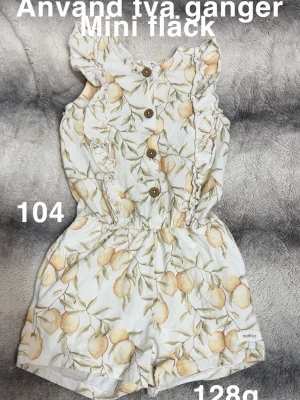 Vit jumpsuit med citronmönster - Supersöt vit jumpsuit från Newbie med citronmönster och volangdetaljer längs axlarna. Knappar framtill och resår i midjan för en bekväm passform. Perfekt för varma dagar och ger en fräsch sommarkänsla.🌸 Använd två gånger. Mini fläck. Storlek 104