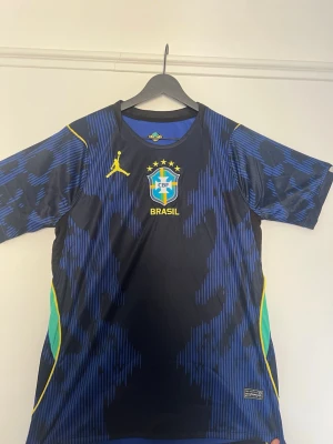Brasilien vm t-shirt Jordan - Snygg svart och blå vm t-shirt från Jordan x Brasilien med CBF-logga och fem stjärnor på bröstet. Gula detaljer och gröna inslag på sidorna. Tillverkad i lätt och ventilerande material, perfekt för fotbollsträning.