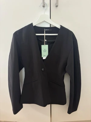 Svart kavaj från Gina Tricot - Stilren svart kavaj från Gina Tricot med djup V-ringning och enkelknäppning. Kavajen har två fickor framtill och en figurnära passform. Perfekt för en trendig och clean look. Materialet är slätt och känns bekvämt mot huden. Helt ny!
