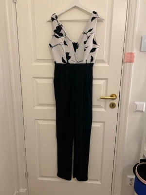 Svartvit blommig jumpsuit med V-ringning - Snygg jumpsuit med svartvita kontraster, överdel i vitt med svart blommönster och djup V-ringning. Nedre delen är helsvart med raka ben. Perfekt för dig som vill sticka ut med stilren look. Materialet känns mjukt och följsamt.