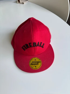 Röd snapback keps FIREBALL - Riktigt snygg röd snapback keps med svart broderad text 'FIREBALL' framtill. Kepsen har platt skärm och justerbar snapback baktill. Perfekt för dig som vill sticka ut med en färgstark accessoar.