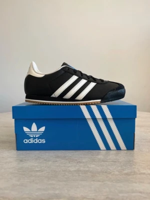 Adidas svartvita sneakers - Klassiska Adidas sneakers i svart med vita ränder och vit häl. Skorna har låg profil, svart ovandel i syntet och mockadetaljer, samt gummisula i brunt. Perfekt för dig som gillar retrostil och sportig vibe.