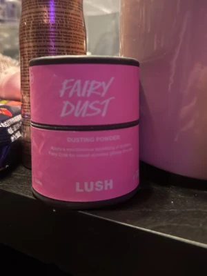 Lush Fairy Dust Dusting Powder - Fairy Dust Dusting Powder från Lush i en rosa burk. Pudret ger en lätt glittrig finish och har en söt doft. Perfekt för att ge huden en mjuk och skimrande känsla. Kommer i en rund förpackning med tydlig text.