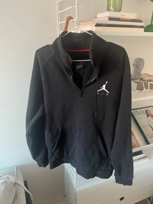 Svart Jordan hoodie med halv dragkedja - Svart hoodie från Jordan med halv dragkedja och vit Jumpman-logga på bröstet. Hoodien har känguruficka, ribbade muddar och mjukt material på insidan. Perfekt för en sportig och avslappnad stil.