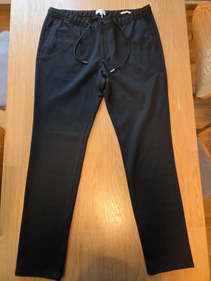Jules svarta slim chinos byxor, storlek 46 - Eleganta svarta byxor med dragsko i midjan. Byxorna har en smal passform och praktiska fickor både fram och bak. Perfekt för en stilren och bekväm look.