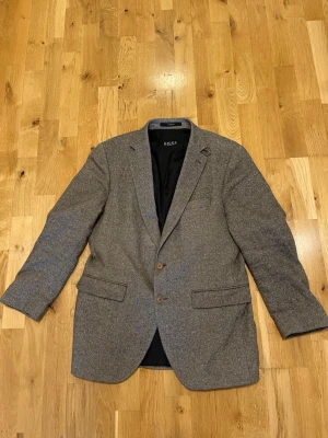 Grå/brun  kavaj från Digel med två knappar stl 48 - Stilren brun/grå kavaj från Digel med klassisk passform och två knappar framtill. Kavajen har två fickor med lock och en bröstficka. Materialet är troligtvis ullblandning och den har svart innerfoder. Perfekt för att höja din outfit.