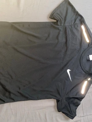 Svart Nike Miller t shirt - Svart tränings-tshirt från Nike med vit logga på bröstet och reflekterande detaljer på axlarna. Tillverkad i ett lätt och snabbtorkande material som passar perfekt för löpning eller gym. Klassisk rund hals och korta ärmar.