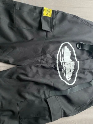 Svarta cargobyxor från corteiz - Svarta cargobyxor från corteiz med stort vitt tryck på benet och flera praktiska fickor. Byxorna har en cool streetwear-vibe och är tillverkade i slitstarkt material. Perfekta för dig som gillar en avslappnad och edgy stil.
