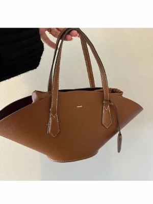 Brun handväska i läderlook - Snygg brun handväska med unik, geometrisk form och dubbla handtag. Väskan har vita kontrastsömmar och en liten dekorativ detalj framtill. Tillverkad i ett material som liknar läder och har en mjuk, strukturerad yta. Perfekt för dig som vill sticka ut med din stil.