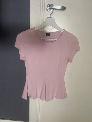 Ljusrosa topp från Gina Tricot - Söt ljusrosa topp från Gina Tricot med rund halsringning och korta ärmar. Modellen är figurnära med lätt utsvängd nederkant, perfekt för en avslappnad och trendig look.