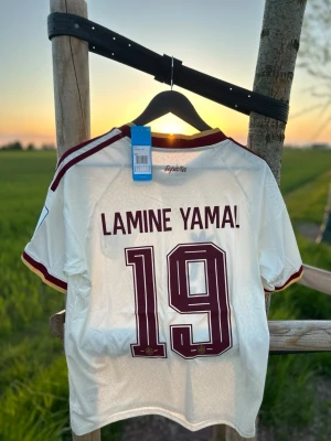 Vit Spanien tröja Lamine Yamal - Säljer en vit Spanien fotbollströja från Adidas med Lamine Yamal och nummer 19 på ryggen. Tröjan har vinröda detaljer, tre ränder på axlarna och officiellt landslagsmärke framtill. 