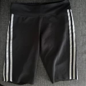 Svarta cykelshorts från Adidas - Svarta cykelshorts från Adidas med klassiska vita ränder längs sidorna. Shortsens material är stretchigt och klippta perfekt för träning. 