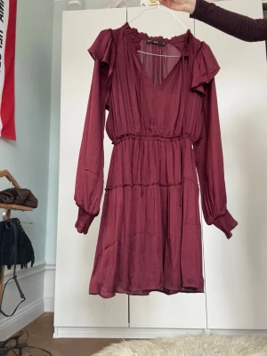 Vinröd maje liknande klänning - Vinröd kortklänning med långa ärmar, v-ringning och volangdetaljer vid axlar och hals. Klänningen har omlottkänsla och markerad midja med resår. Tillverkad i ett lätt, flowy material som ger en luftig känsla.
