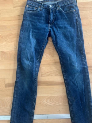 Mörkblå jeans med raka ben - Säljer ett par klassiska mörkblå jeans med raka ben och femficksdesign. Jeansen har en knappstängning och subtila slitningar som ger en cool look. Perfekta för dig som gillar tidlös stil och denim.