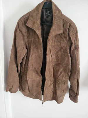 Brun skinnjacka från Heritage - Snygg brun skinnjacka från Heritage i klassisk modell med krage och dragkedja framtill. Jackan har två fickor på sidorna och är tillverkad i mjukt skinn som ger en cool vintagekänsla. Perfekt för dig som vill ha en tidlös och stilren look.