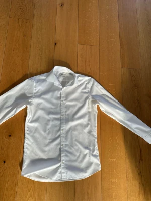 Vit slim fit skjorta Jack & Jones - Klassisk vit skjorta från Jack & Jones i slim fit-modell. Skjortan har långa ärmar, knappar framtill och en stilren krage. Tillverkad i bomull för en skön känsla och snygg passform. Perfekt för dig som gillar en clean och enkel look.