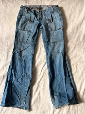 Blå bootcut jeans från Mavi - Säljer ett par ljusblå bootcut jeans från Mavi med coola detaljer som stora fickor framtill, dekorativa sömmar och dragkedja. Jeansen har en avslappnad passform och är tillverkade i mjukt denimtyg. Perfekta för en casual och trendig look.